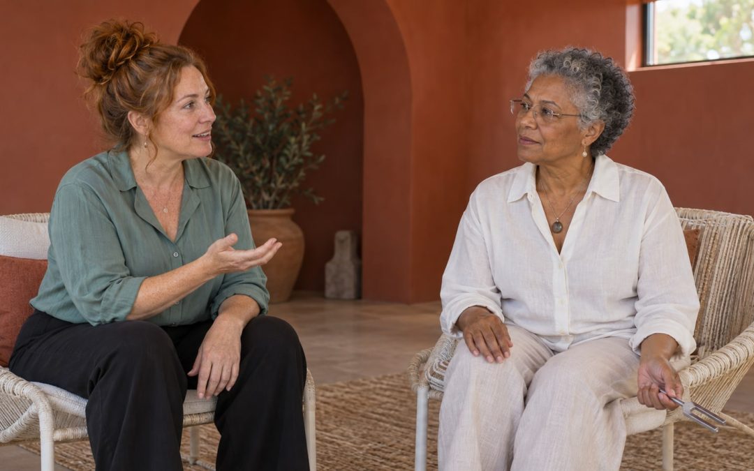 Two women in their fifties and sixties talking in a quiet room, with one holding a small tuning fork away from both people.