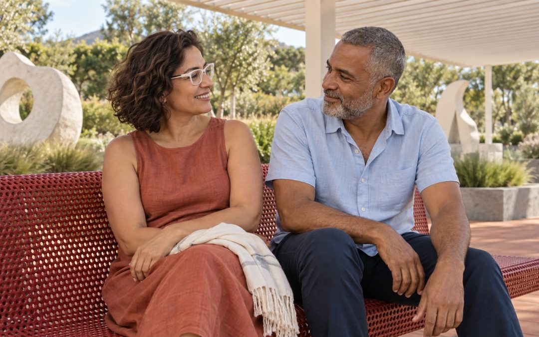 A mature woman in white glasses and a terracotta dress talks with a gray-haired bearded man on a red metal bench in a bright sculpture garden.